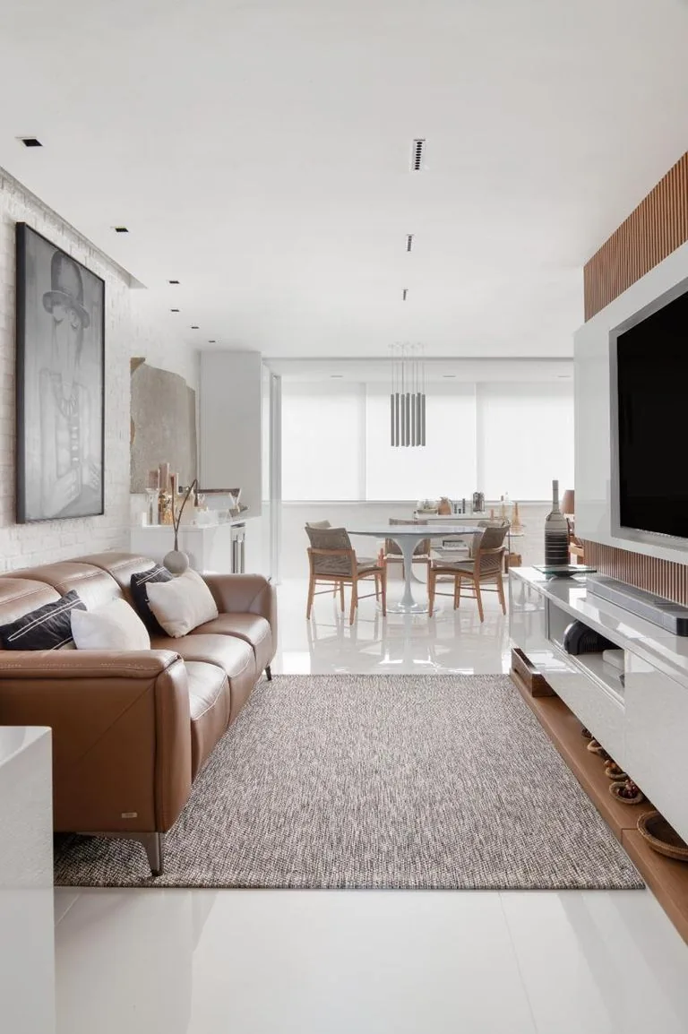 Living em tons claros com sofá de couro caramelo, tapete e painel de TV, com a sala de jantar ao fundo