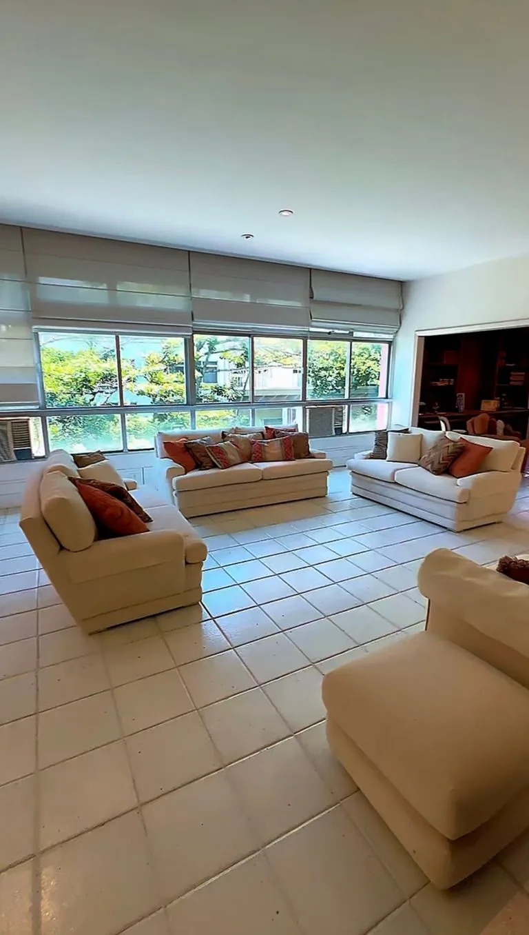 Sala ampla com três sofás creme e almofadas coloridas, piso de cerâmica clara e janelas com persianas